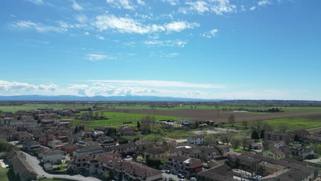 Drone-Vuela-Sobre-Una-Ciudad-En-El-Valle-Del-Po-Con-Los-Alpes-Al-Fondo-En-Un-Soleado-Día-De-Primavera