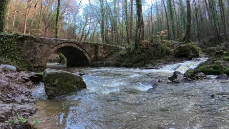 Fluss-Fließt-Aus-Einem-Kleinen-Waldwasserfall-Unter-Einer-Rustikalen-Steinfußgängerbrücke-Im-Herbstwald-In-Zeitlupe