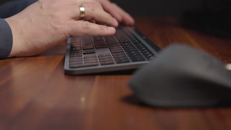 Person’s-hands-wearing-a-grey-sweater-and-has-a-ring-on-the-left-handtyping-on-a-black-keyboard-placed-on-a-polished-wooden-desk
