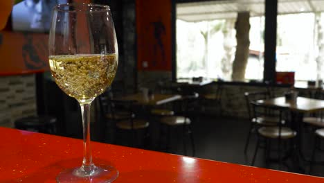 Leeres-Glas-Wird-Mit-Weißem-Wein-Gefüllt,-Während-Im-Hintergrund-Des-Leeren-Restaurants-Ein-Fernseher-Läuft
