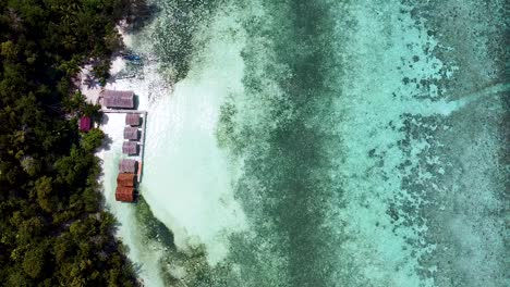 Luftaufnahme-Abgelegener-Strandhütten-Mit-Blick-Auf-Atemberaubendes-Kristallklares-Meerwasser-Und-Korallenriffe-Auf-Einer-Exotischen,-Abgelegenen-Tropischen-Insel-In-Raja-Ampat,-West-Papua,-Indonesien