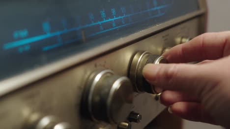 Die-Hand-Einer-Person,-Die-Knöpfe-Dreht,-Um-Den-Sender-Eines-Vintage-Radios-Mit-Metallischen-Knöpfen-Und-Einem-Blau-Beleuchteten-Analogen-Frequenzrad-Einzustellen
