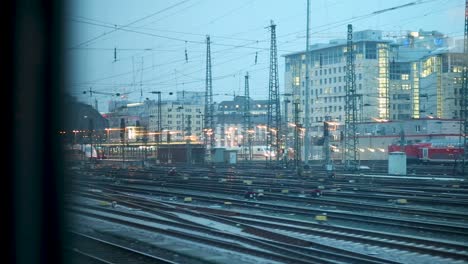 Blick-Aus-Einem-Zug-Auf-Belebte-Bahngleise-Und-Städtische-Gebäude-In-Der-Abenddämmerung