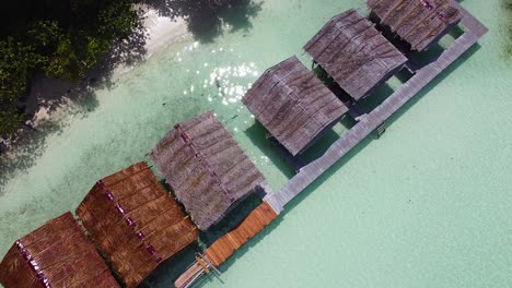 Static-aerial-view-above-remote,-rustic-huts-overlooking-crystal-clear-ocean-water-on-tropical-island-in-Raja-Ampat,-West-Papua,-Indonesia
