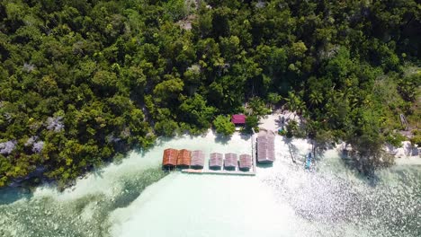 Aerial-drone-rising-above-a-row-of-waterfront-beach-huts-overlooking-ocean-on-rainforest-covered-island-in-Raja-Ampat,-West-Papua,-Indonesia