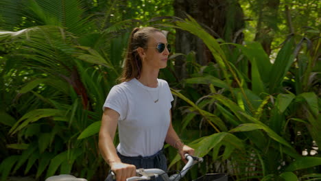 Young-female-traveler-with-sunglasses-walking-through-palm-trees-with-her-bike-to-the-beach