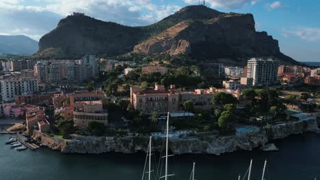 Drone-flying-backwards-in-Palermo's-bay-area-while-hotels,-sailing-boats-and-blue-sea-visible-in-the-foreground-and-Monte-Pellegrino-mountain-in-the-background