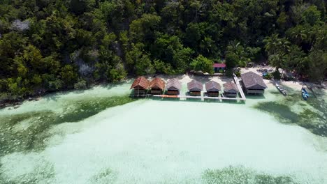 Aerial-pull-out-of-exotic-waterfront-huts-over-crystal-clear-ocean-water-in-Raja-Ampat,-West-Papua,-Indonesia