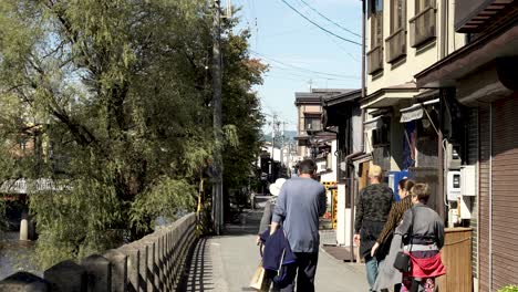 Family-of-tourists-walking-through-an-alley-and-bridge-in-Japan,-surrounded-by-a-large-tree,-a-river-and-houses-with-ancient-architecture