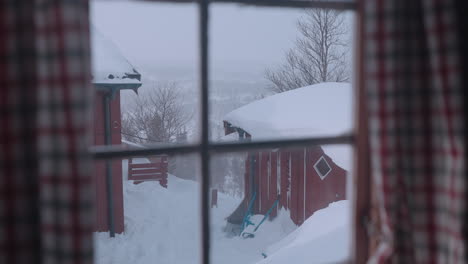 Cozy-view-from-a-Norwegian-cabin-window,-capturing-a-serene-winter-forest-scene-with-gently-falling-snow-and-charming-red-cabins-nestled-among-snow-covered-trees