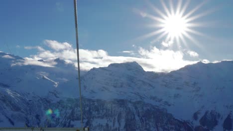 View-from-above-of-a-single-cable-used-for-a-number-of-rides-going-up-and-down-the-slopes,-showing-the-glacial-mountains,-sun-,-and-blue-cloudy-skies-on-the-other-side,-at-Engelberg,-Switzerland