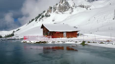 Dron-Panorámico-Desde-El-Lado-Derecho-Al-Izquierdo-Del-Cuadro,-Mostrando-Un-Lago-Congelado-Frente-A-Una-Cabaña-De-Madera-En-Una-Ladera-Nevada-De-Los-Alpes-En-Engelberg,-Suiza