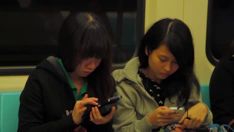 Dos-Jóvenes-Adultas-Asiáticas-Ocupadas-Con-Sus-Teléfonos-En-El-Metro