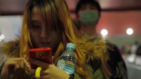 Asiatisches-Mädchen-Mit-Rotem-Telefon-Und-Einer-Flasche-Wasser-In-Der-Hand,-Das-Auf-Der-Rolltreppe-Tippt