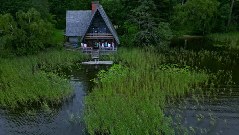 Aerial-drone-backward-moving-shot-over-family-and-friends-enjoying-at-a-wooden-cottage-by-the-shore-of-lake-surrounded-by-lush-green-vegetation-at-daytime