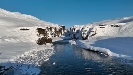 Vista-Aérea-Sobre-El-Agua-De-Un-Río-Glaciar-Que-Fluye-A-Través-De-Un-Cañón-Cubierto-De-Nieve,-En-Un-Día-Soleado