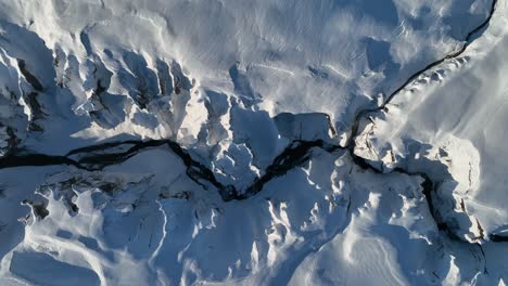 Aerial-top-view-of-a-glacier-river-flowing-through-a-canyon-covered-in-snow,-on-a-sunny-day