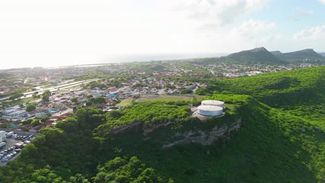 Large-water-tanks-sit-on-tall-tropical-hill-overlooking-Caribbean-island,-aerial-establish