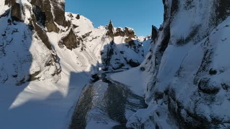 Aerial-view-through-a-canyon-covered-in-snow,-over-a-glacier-river-flowing,-on-a-sunny-day