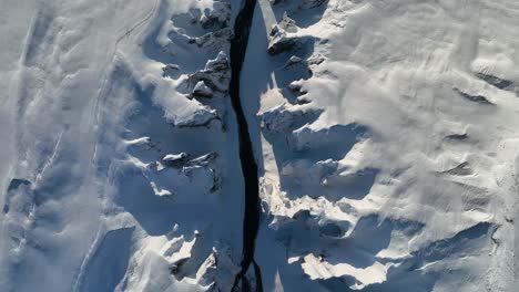 Aerial-top-view-over-a-glacier-river-flowing-through-a-snow-capped-canyon,-on-a-sunny-day