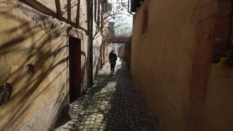 Female-tourist-walking-around-sunny-small-street-with-cobblestone-roads-and-half-timbered-houses-in-Kaysersberg,-France