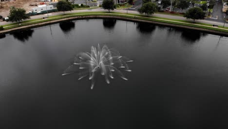Aerial-panning-shot-around-fountain-in-pond-reflections-in-the-water-with-trees-in-the-background