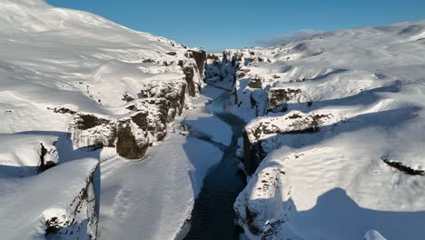 Aerial-view-of-a-glacier-river-flowing-through-a-canyon-covered-in-snow,-on-a-sunny-day