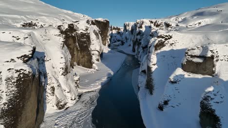 Aerial-view-over-a-glacier-river-flowing-through-a-canyon-covered-in-snow,-on-a-sunny-day