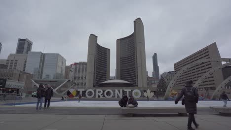 Aufnahme-Des-Nathan-Phillips-Square-Und-Des-Rathauses-Von-Toronto-Mit-Ein-Paar-Jungen-Beim-Schlittschuhlaufen-Und-Leuten,-Die-An-Einem-Kalten,-Wolkigen-Abend-In-Toronto,-Kanada,-Durch-Das-Rathaus-Laufen