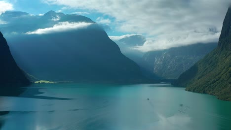 Un-Vuelo-Sobre-Las-Aguas-Turquesas-Del-Lago-Noruego-Loenvatnet-Con-Montañas-Que-Se-Elevan-A-Los-Lados-Raspando-Las-Nubes-Blancas-Bajas