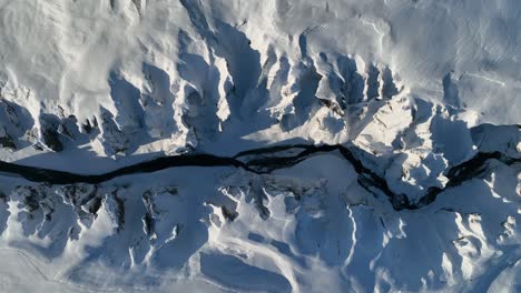 Aerial-top-view-over-a-glacier-river-flowing-through-a-canyon-covered-in-snow,-on-a-sunny-day