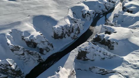 Aerial-view-over-a-glacier-river-flowing-through-a-canyon-covered-in-snow,-on-a-sunny-evening