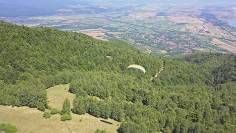 Scenic-view-following-paraglider-over-green-trees-with-valley-and-town-at-base-of-mountain