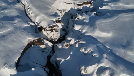 Aerial-landscape-view-over-a-glacier-river-flowing-through-a-canyon-covered-in-snow,-on-a-sunny-day