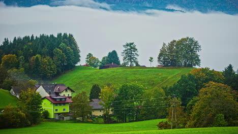 Cozy-Alpine-village-with-fog-flowing-in-above-clouds,-time-lapse-view