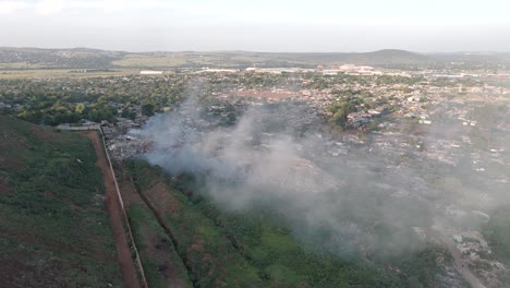 Captivating-4K-aerial-footage-features-a-dynamic-push-in-drone-shot,-seamlessly-revealing-a-rural-township-village-situated-on-the-edge-of-a-waste-processing-plant-burning-waste-in-South-Africa