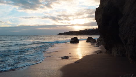 Pov-walk-through-hole-of-cliff-reaching-sandy-beach-with-waves-at-Algarve-at-sunset-time