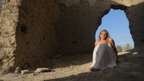 A-woman-in-a-white-dress-sits-as-the-base-of-ancient-ruins-with-the-wind-blowing-on-a-sunny-day