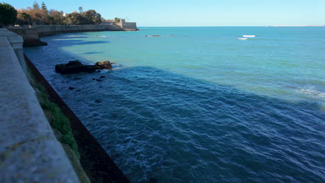 Tranquilas-Aguas-Azules-Junto-A-Un-Malecón-De-Piedra,-Con-Vistas-A-La-Costa-Lejana