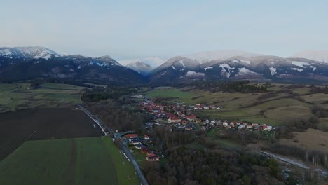 Fly-above-small-village-with-houses-and-snow-capped-mountains-in-the-background