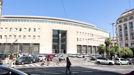 Un-Edificio-Moderno-En-Nápoles-Con-Una-Fachada-Curva,-Grandes-Ventanas-Verticales-Y-Banderas-Italianas,-Yuxtapuesto-A-Una-Escena-Callejera-Muy-Transitada-Con-Peatones-Y-Vehículos.