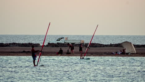 Windsurfistas-Preparando-Su-Equipo-Al-Atardecer-En-Un-Mar-Tranquilo-Con-Fondo-De-Playa