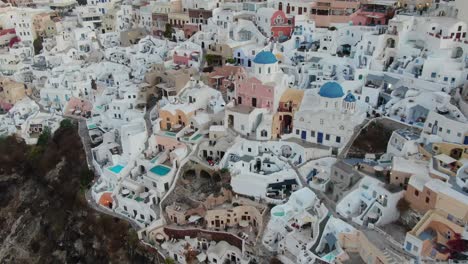 Drone-view-in-Greece-flying-over-Santorini-with-Oia-town-white-houses-and-blue-roofs-on-a-cliff-next-to-the-mediterranean-sea-at-sunrise