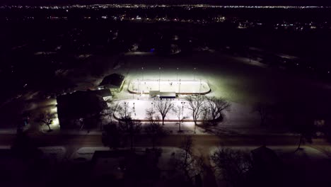 Canadian-winter-night-scene,-outdoor-hockey-rinks-illuminated-by-floodlight-and-city-skyline,-4K-drone-shot