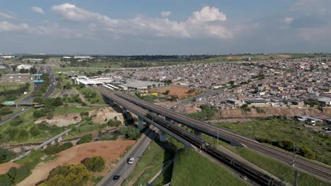 4K-aerial-clip,-a-static-drone-captures-the-dynamic-energy-of-a-high-speed-train,-juxtaposed-against-the-bustling-highways-and-the-expansive-urban-township-of-Alexandra-in-Johannesburg,-South-Africa