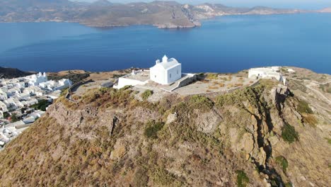 Drone-view-in-Greece-circling-around-a-white-church-at-the-top-of-a-brown-hill-with-a-greek-white-house-town-facing-blue-sea-on-a-mountain