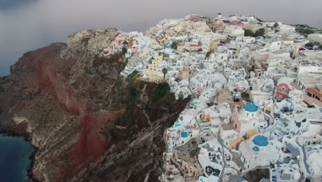 Drohnenansicht-In-Griechenland,-Die-Bei-Sonnenaufgang-über-Santorini-Mit-Weißen-Häusern-Und-Blauen-Dächern-Der-Stadt-Oia-Auf-Einer-Klippe-Am-Mittelmeer-Fliegt