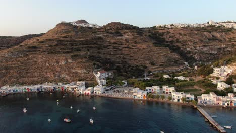 Drohnenansicht-In-Griechenland,-Vertikaler-Aufstieg-über-Eine-Kleine-Stadt-Mit-Weißen-Häusern-Und-Farbigen-Türen-Neben-Dem-Meer-Vor-Einem-Berg-In-Milos-Bei-Sonnenuntergang