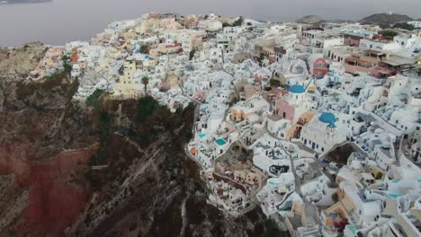 Drone-view-in-Greece-flying-over-Santorini-with-Oia-town-white-houses-and-blue-roofs-on-a-cliff-next-to-the-mediterranean-sea-at-sunrise