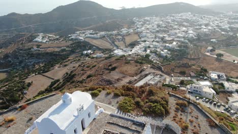 Drone-view-in-Greece-flying-over-a-small-white-church-at-the-top-of-a-brown-hill-with-a-greek-white-house-town-facing-blue-sea-on-a-mountain